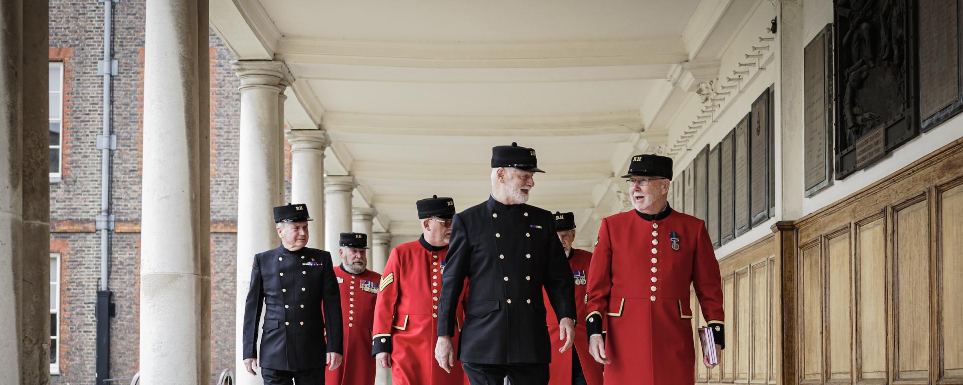 A group of Chelsea Pensioners dress in a mix of iconic scarlet and blues uniform, walk together happily along a wood panelled colonnade.