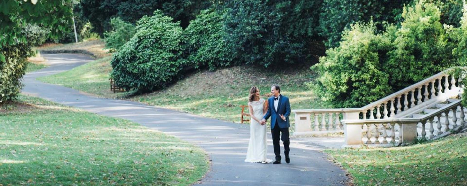 Bride and groom walking hand in hand through the grounds of the Royal Hospital Chelsea, with a stone staircase and mature trees in the background.