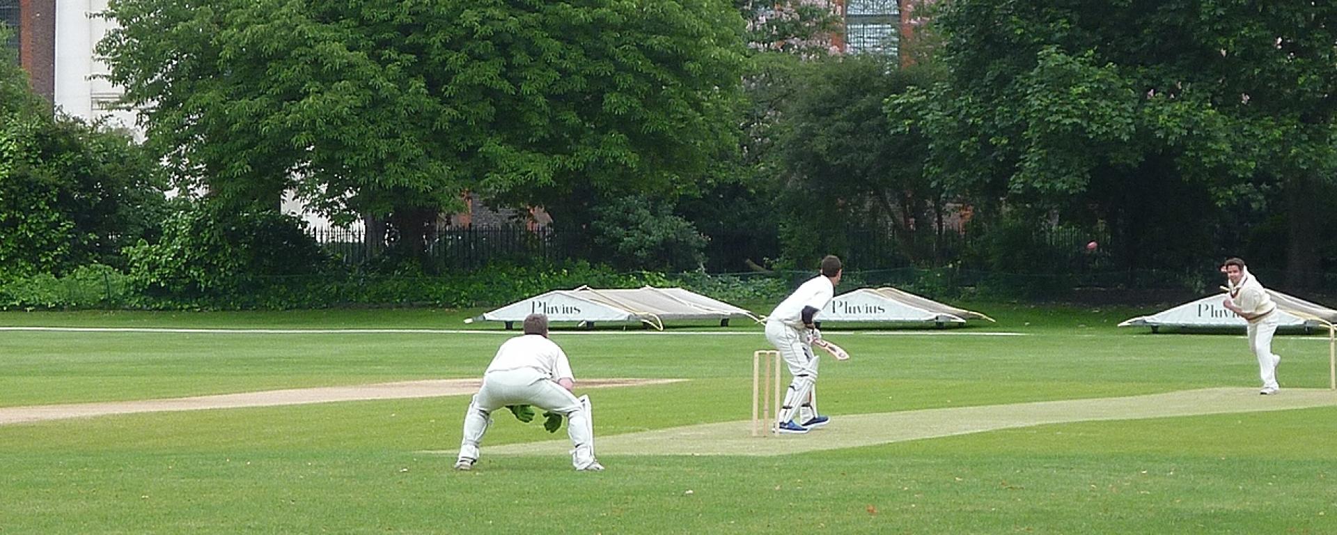 Players competing in a cricket match on the Royal Hospital Chelsea’s historic cricket ground, surrounded by mature trees and the Hospital’s heritage buildings.