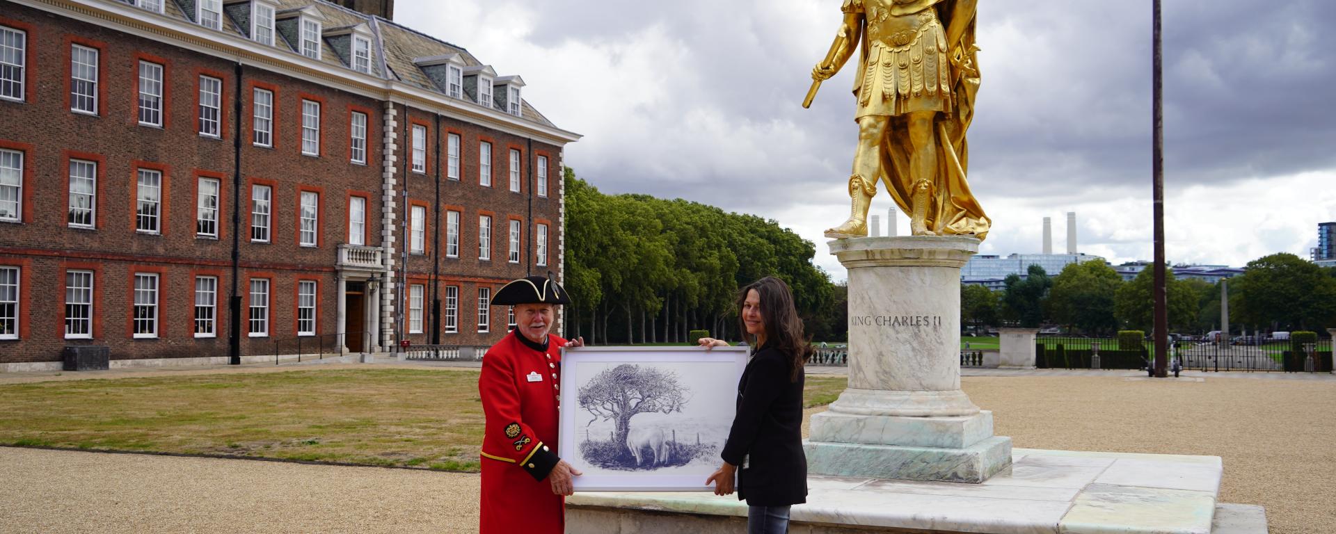 An image of Chelsea Pensioner Roy Palmer holding up a painting alongside a woman with a gold statue in the background at Royal Hospital Chelsea