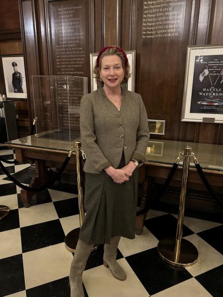 A lady, dressed in a tweed blazer, dark long skirt and grey boots, stands on a checkered black and white floor in the Great Hall at The Royal Hospital Chelsea. Wood panels adorn the walls and a large wooden table with a glass case can be seen behind her.