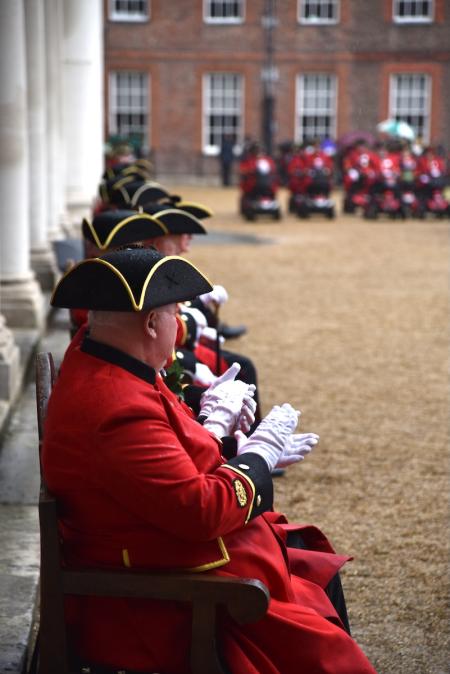 Chelsea Pensioners sit along a gravel parade ground, dressed in their iconic scarlet uniforms and tricorn hats and white gloves.