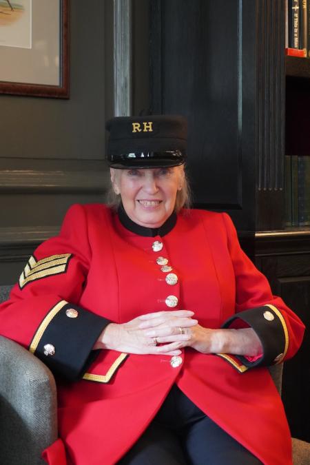 A lady Chelsea Pensioner is sat in her signature scarlet uniform and black shako hat. Within a wood panelled room.