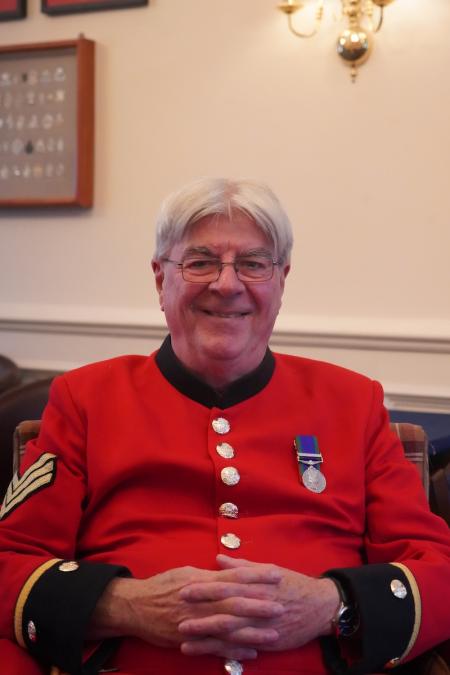 A male Chelsea Pensioner sits on a brown armchair, dressed in iconic scarlet uniform and glasses. A single medal adorn's his left breast.