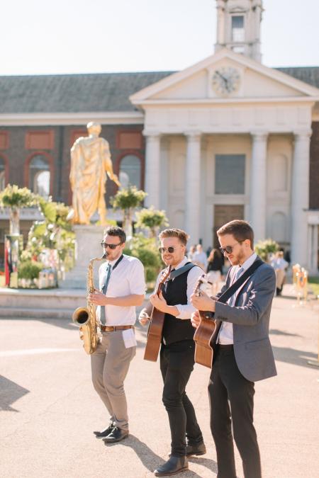 Three musicians perform outdoors in bright sunlight, standing in front of the Royal Hospital Chelsea. One plays a saxophone and the other two play acoustic guitars. All three wear sunglasses and smart attire. A large golden statue and the building’s classical facade with columns and a clock tower are visible in the background.