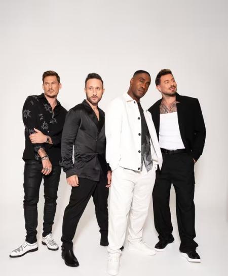 Four male performers pose together against a plain light background. They wear coordinated modern outfits in black and white, standing close and facing the camera with relaxed, confident expressions, suggesting a professional band or vocal group promotional image.