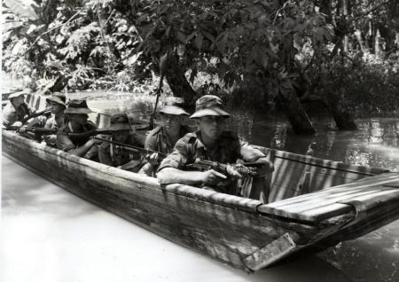 A historic black and white photo of Borneo, Indonesia depicting a boat patrol with multiple military figures in camouflage uniform and weapons drawn