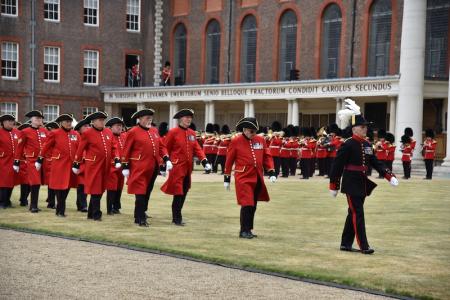 Chelsea Pensioners in scarlet uniforms march in formation across the lawn at the Royal Hospital Chelsea, led by an officer in black ceremonial dress. A military band performs behind them, with the historic colonnade and brick buildings in the background.