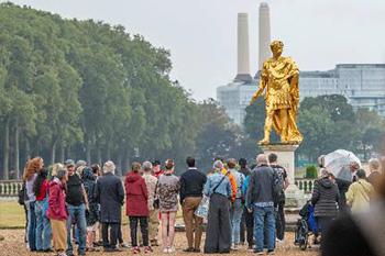 A group of people gather around a gold statue of King Charles II in the south grounds of The Royal Hospital Chelsea. Green trees flank the grounds to the left and battersea power station's famous chimneys can be see in the background.