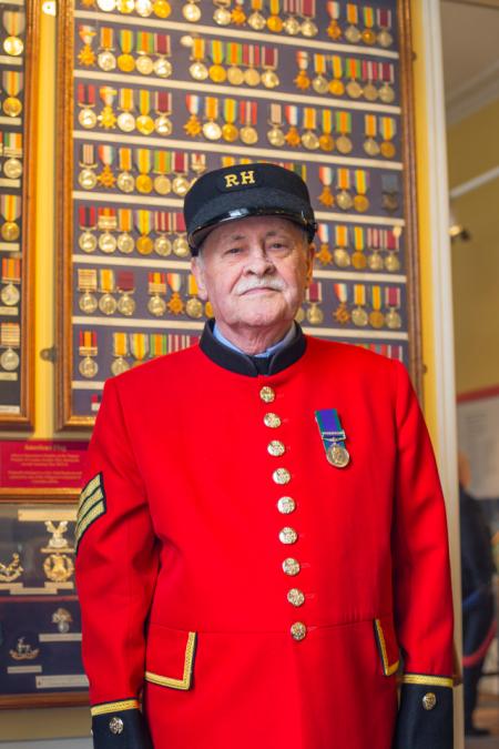 A Chelsea Pensioner stands in front of a wall of medals, wearing his iconic scarlet uniform adorned with a single medal on the lapel.