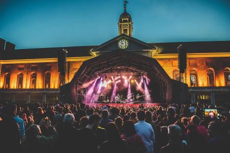 A large outdoor concert at dusk takes place in front of a historic building with a central clock tower. A covered stage is lit with purple and white lights as a band performs, while a large crowd fills the foreground, watching the show against the warmly illuminated brick façade.