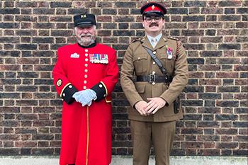 A Chelsea Pensioner, dressed in his iconic scarlet uniform adorned with medals, and wearing a black Shakko hat. Besides him stands are younger serving military personnel in traditional khaki coloured uniform and a black brimmed hat with red accent