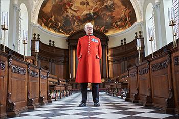 A Chelsea Pensioner, dressed in his iconic scarlet uniform, stands within a grand chapel with dark wooden pews on either side of a chequered black and white aisle. Leading to a wooden altar with a painted fresco above