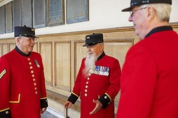 Three Chelsea Pensioners in traditional scarlet uniforms stand together indoors, talking and smiling. They wear black RH caps and service medals. The setting features wooden wall panelling and a light, historic interior at the Royal Hospital Chelsea.