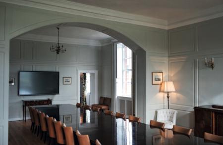 A stately boardroom with classic panelled walls painted in soft grey-green tones. A long, polished wooden table is surrounded by tan leather chairs. The room features a flat-screen TV, traditional chandeliers, a standing lamp beside a white tufted armchair, and framed artwork on the walls. Natural light streams through tall sash windows, creating a blend of heritage charm and modern functionality.