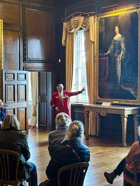 Chelsea Pensioner in a scarlet uniform leading a guided tour, pointing to a large portrait painting inside a historic wood-panelled room at the Royal Hospital Chelsea.