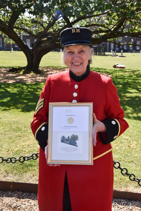 Chelsea Pensioner Barbara, in scarlet uniform holds a framed RHS gold medal certificate in the grounds of the Royal Hospital Chelsea.
