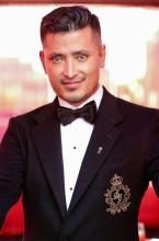 A man wearing a black tuxedo jacket with a bow tie and an embroidered crest on the breast pocket looks towards the camera. He has neatly styled dark hair and stands against a softly lit background with warm tones, suggesting a formal or celebratory event setting.