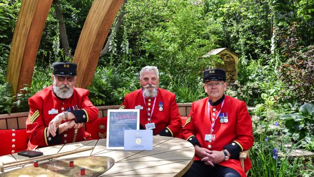 Chelsea Pensioners sit behind a round wooden table which displays their Gold Award from the Chelsea Flower Show 2025