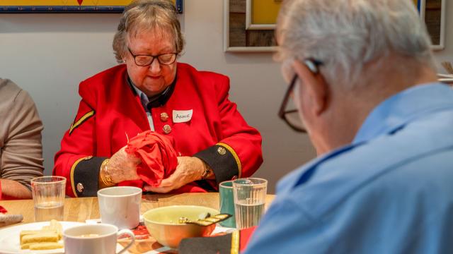 An older woman wearing the iconic red Chelsea Pensioners uniform, identified by a name tag as “Anne”, sits at a table engaged in hand-sewing. She is carefully mending a red garment, surrounded by cups, plates of biscuits, and a warm, communal atmosphere.