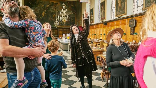 An actor dressed in period costume and representing Sir Christopher Wren stands inside the Great Hall at the Royal Hospital Chelsea with wood-panelled walls and chequered black and white floor tiles. A group of visitors of various ages listen in
