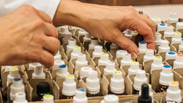 Hands selecting from a number of small vials with white lids. A series of small drawers in the background.