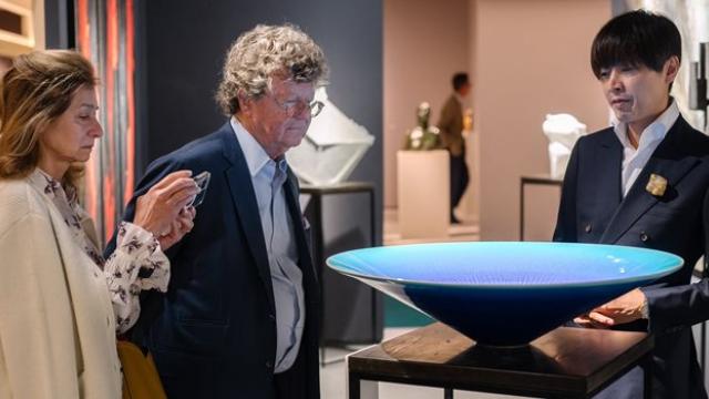 Two men and a lady stand behind a giant coral blue vase balanced upon a wooden plinth