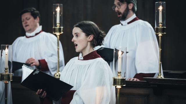 Two male and one female sing in traditional white church robes with maroon collars and sleeves indoors by candlelight