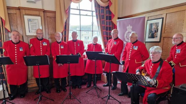A group of scarlet clad Chelsea Pensioners are arranged behind a series of music stands in a wood panelled room with dark red carpet
