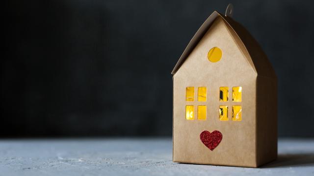 A hand crafter cardboard house sits on a blank grey surface in front of a dark black background, contrasted by the glowing yellow light shining through the windows of the house and accented with a red heart below the windows.