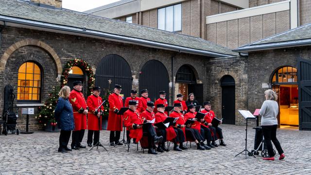 Two rows of Chelsea Pensioners, the front row seated and back row standing, wear their iconic scarlet uniforms as they sign in a stone courtyard, decorated with green, red and gold Christmas trees and garlands