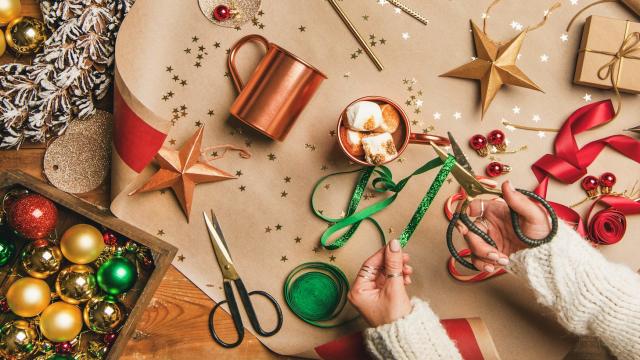 A colourful table top is shown from above with red, yellow and green decorations and crafting equipment. A pair of hands are crafting decorations while a mug of hot chocolate with huge fluffy white marshmallows sits on the table.