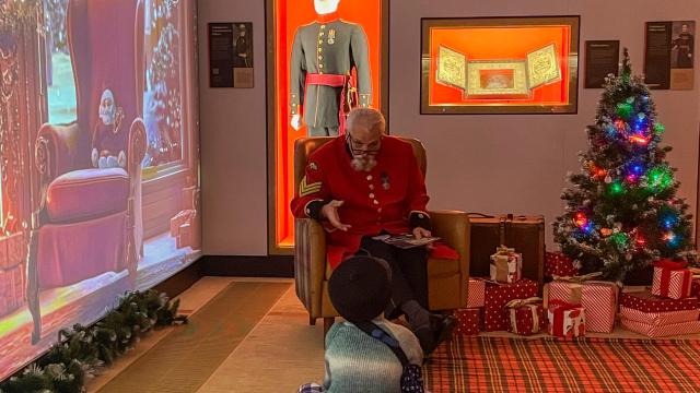 A Chelsea Pensioners sits in front of a young child in a Christmas themed grotto. A Christmas tree and lights twinkle in the corner and a digital backdrop extends the theme.