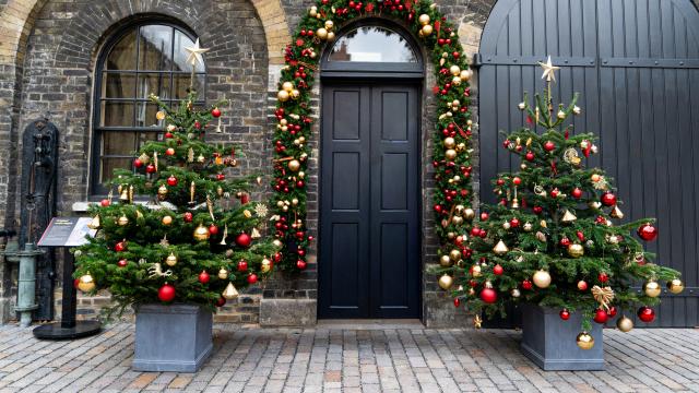 Two beautifully decorated gold, silver and red Christmas Trees stand in an outdoor courtyard besides a big black archway door with a festival garland surrounding. 