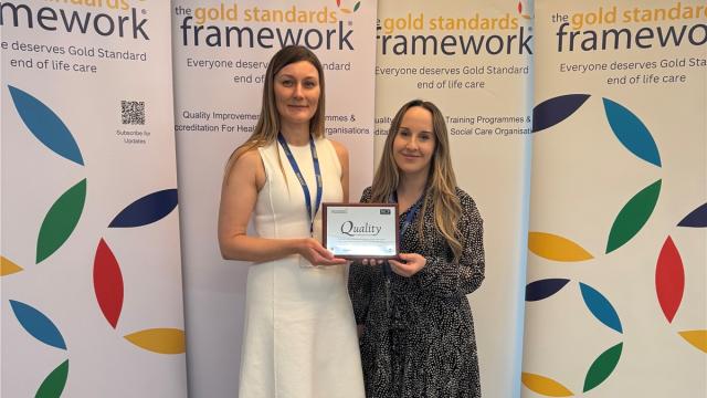 Royal Hospital Chelsea's Matron dressed in a long while dress, and Deputy Matron in a dark pattern dress, stand in front of a coloured Gold Standards Framework banner holding an award for Outstanding End of Life Care