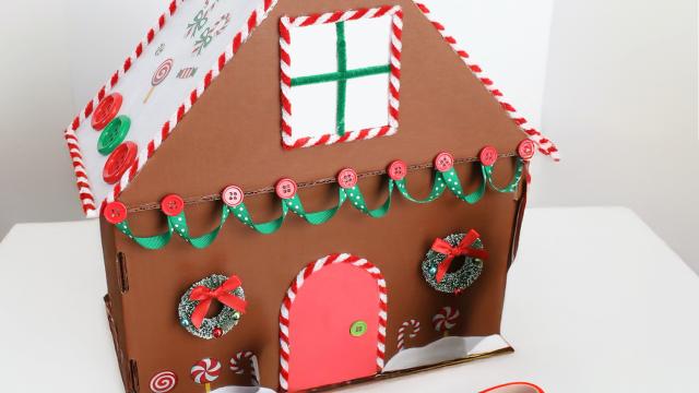 A cardboard gingerbread house sits atop a white table with colourful Christmas buttons and ribbons decorating the house. On the table are strewn buttons of various colours and a red pair of scissors