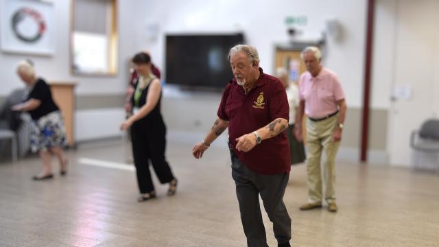 A retired older man stands at the front of a class of other adults, with a headset, instructing them in dancing.