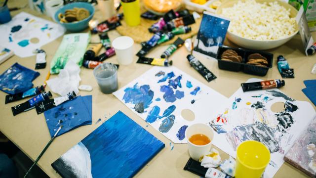 A table scattered with art suppliers, including paints, canvases, paintbrushes. Blue colours dominate, a glass of yellow liquid and a bowl of popcorn also sit upon the table.