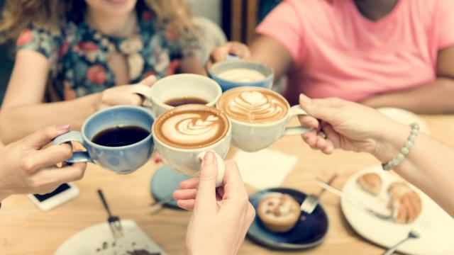 A group of people hold various mugs and cups full of tea and coffee in a collective "cheers" - beneath the cups is a wooden table with saucers, spoons and various pastries strewn about it.