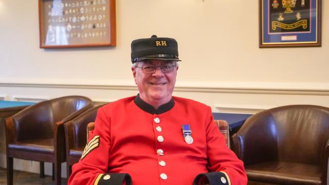 A male Chelsea Pensioner sits on a brown armchair, dressed in iconic scarlet uniform and black shako hat. A single medal adorn's his left breast.