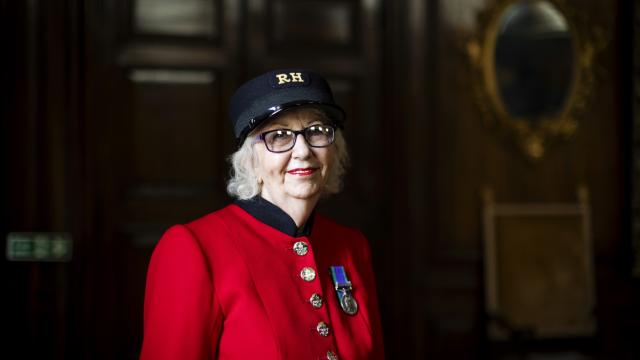 A female Chelsea Pensioner stands wearing her vibrant scarlet uniform, black shako hat and glasses, in front of large wood panelled doors.
