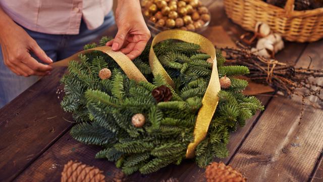 A pair of hands are tying a yellow ribbon on to a handcrafted Christmas wreath of green pine branches. A wooden table holds a container of gold baubles and pine cones.