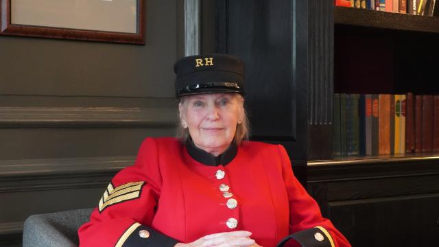 A lady Chelsea Pensioners sits within a wood panelled room in traditional scarlet uniform and black shako hat.