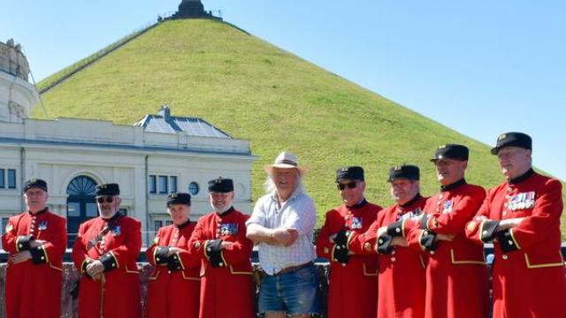 Scarlet clad Chelsea Pensioner stand alongside a civillian in a straw hat. They stand in front of a steep green hill with a monument atop and a white pavilion at the base. A blue clear sky frames the shot.