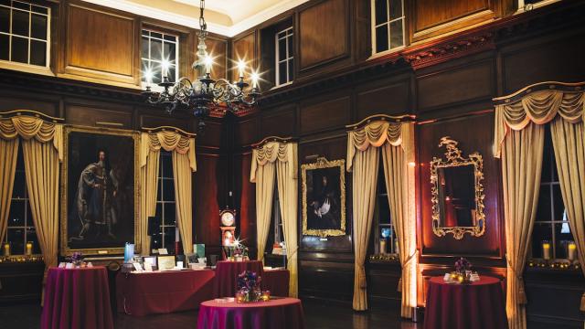 An opulent wood panelled room, with long flowing white curtains and red covered tables throughout the room. Large historic paintings hang on the walls and a warm light emphasises the night time shot.