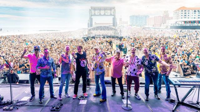 A large band stands across a festival stage facing a vast seaside crowd, smiling and gesturing towards the audience. Thousands of people fill the beach and promenade beyond, with a large outdoor stage structure and coastal buildings visible in the distance under a bright sky.