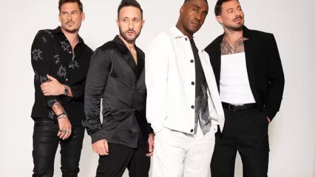 Four male performers pose together against a plain light background. They wear coordinated modern outfits in black and white, standing close and facing the camera with relaxed, confident expressions, suggesting a professional band or vocal group promotional image.