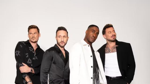 Four male performers pose together against a plain light background. They wear coordinated modern outfits in black and white, standing close and facing the camera with relaxed, confident expressions, suggesting a professional band or vocal group promotional image.