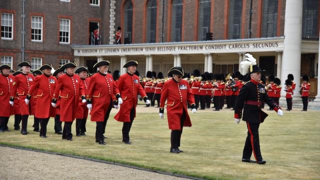 Chelsea Pensioners in scarlet uniforms march in formation across the lawn at the Royal Hospital Chelsea, led by an officer in black ceremonial dress. A military band performs behind them, with the historic colonnade and brick buildings in the background.