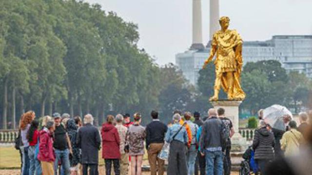 A group of people gather around a gold statue of King Charles II in the south grounds of The Royal Hospital Chelsea. Green trees flank the grounds to the left and battersea power station's famous chimneys can be see in the background.